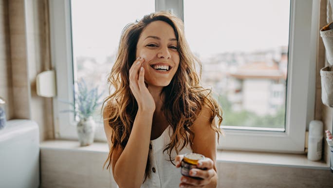 A young woman in front of a mirror puts the best skincare for sensitive skin on her face while smiling.