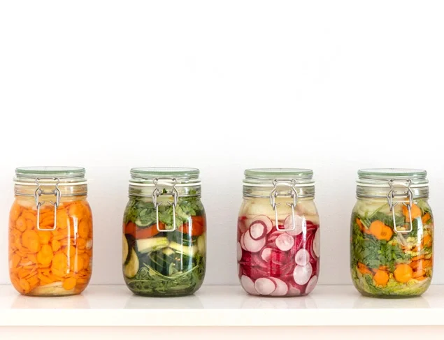 A line of picking jars full of colourful fermented vegetables is displayed in front of a white wall
