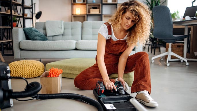 A young woman with curly blonde hair and wearing brown overalls is crouched on the floor assembling a vacuum cleaner