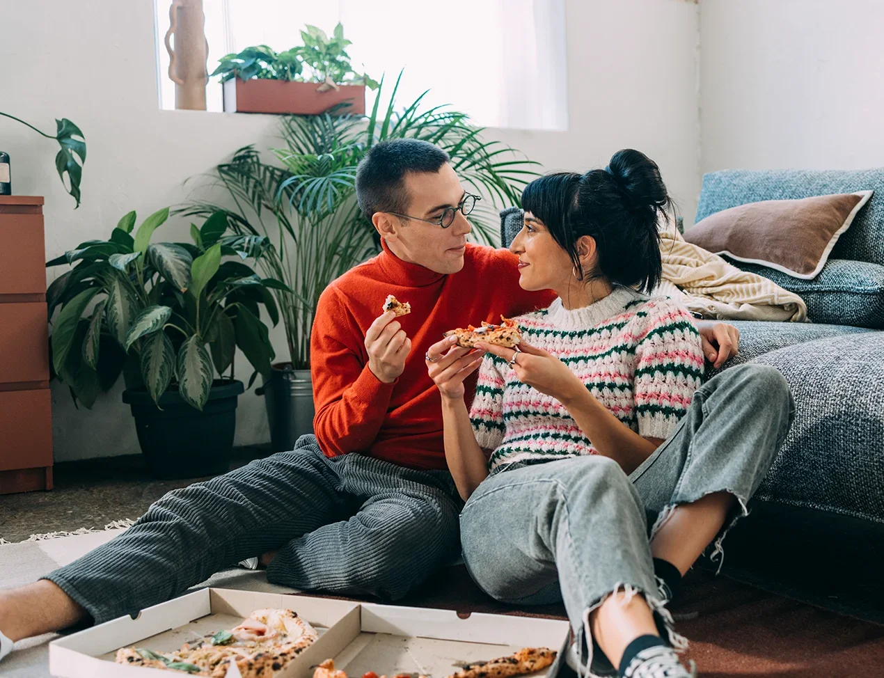 Couple sit on the floor eating pizza together in their living room.
