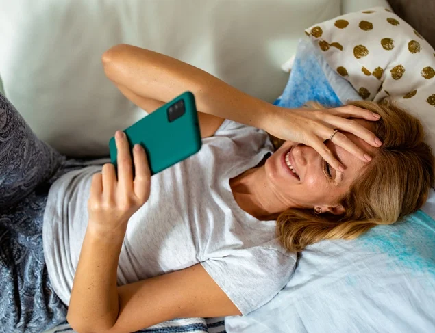 Woman using her smartphone as she lies down the length of the bed and smiling