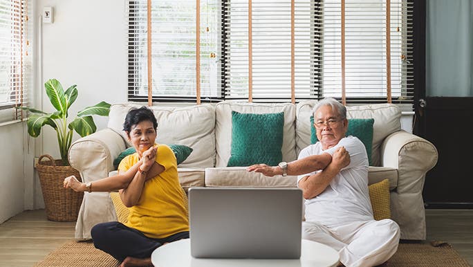  couple sits on the floor in front of a couch. There’s a coffee table with an open laptop in front of them. They look to be doing some sort of guided workout. 
