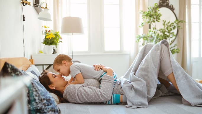 A woman lays in bed with a toddler on her tummy. The age of her child means she can’t get enough sleep.