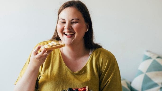 A young woman is smiling as she eats a piece of toast with banana on top