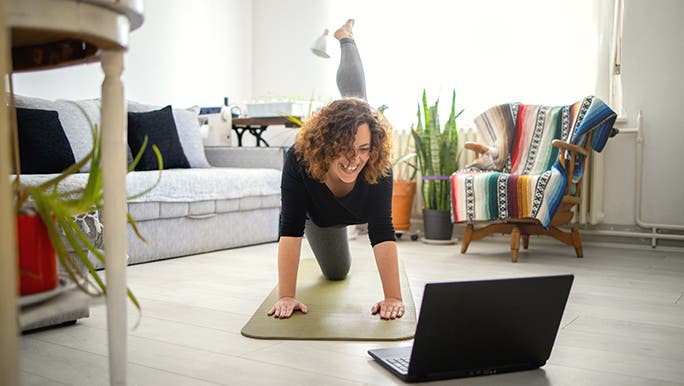 A woman smiles as she works out with her friend via Zoom. She is in a sunny lounge room and it looks like she is doing pilates. 