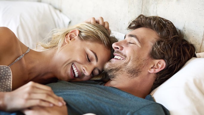 A man and woman are laying on their backs as they’re in bed. They’re cuddling each other and smiling, as she lays her head on his shoulder and one hand on her head. Whilst both holding hands with the other, representing a healthy sex life.