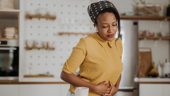 A woman in a kitchen is holding her stomach and looking down at her smartphone while wondering how stress affects the gut. 