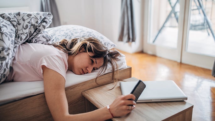 A lady is in her bed, but also on her phone scrolling through social media. Her eyes look tired and stressed, needing a boost of energy before she starts her day.