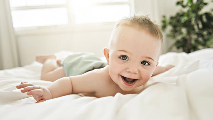  smiling baby is enjoying tummy time on a bed with white sheets. There is a sunny window and a green plant in the background.