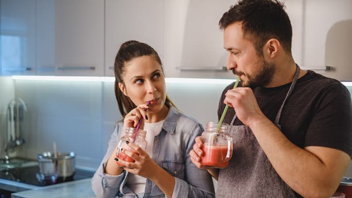 A man and woman drink a smoothie in their kitchen, discussing is it better to eat before or after a workout.