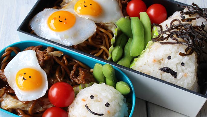 A cute, bento style lunch box with a rice ball, fried egg and noodles.