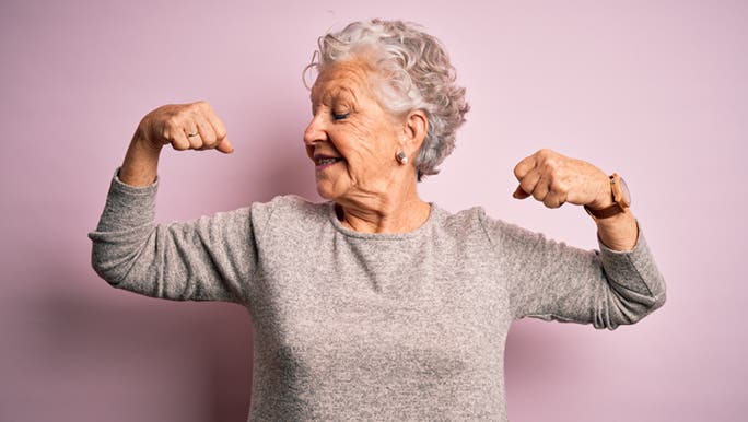 A healthily ageing woman is flexing her muscles and smiling in front of a purple wall. 