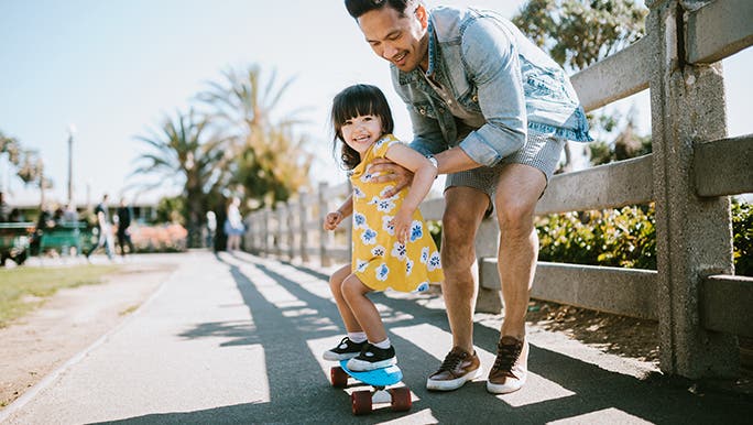 A dad holds his daughter on a skateboard. They are on a footpath that looks like it may be near the beach. 