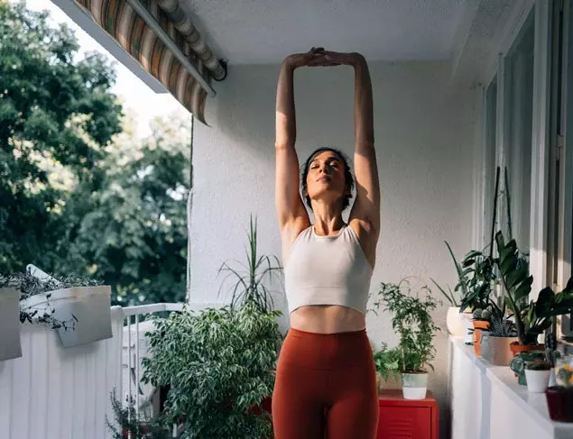 Woman in orange and white active wear standing on her balcony stretching her arms overhead
