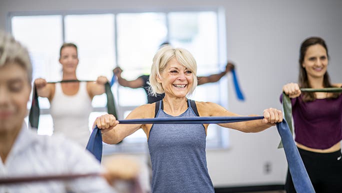 A smiling woman enjoys a group exercise class, she is holding a resistance band in front of her chest. 