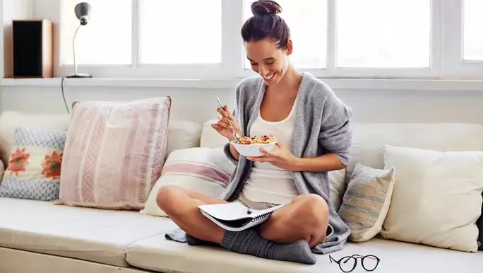 Smiling woman with brown hair eating breakfast in her pyjamas on the couch