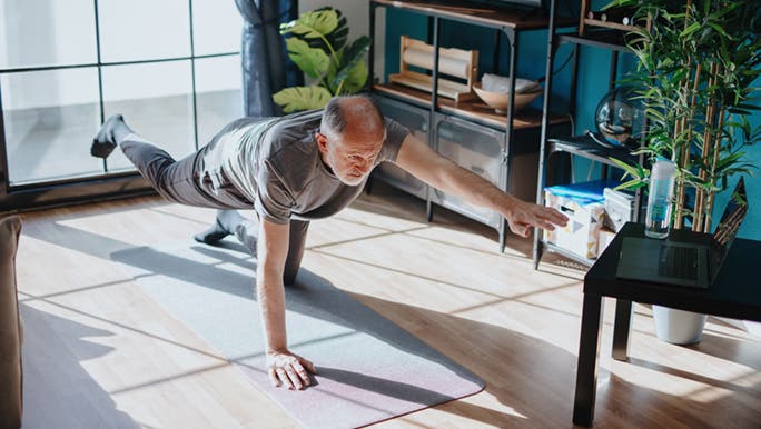 A man balances on one hand and knee on a yoga mat in his home. It seems as though he knows the importance of exercise for older people. 