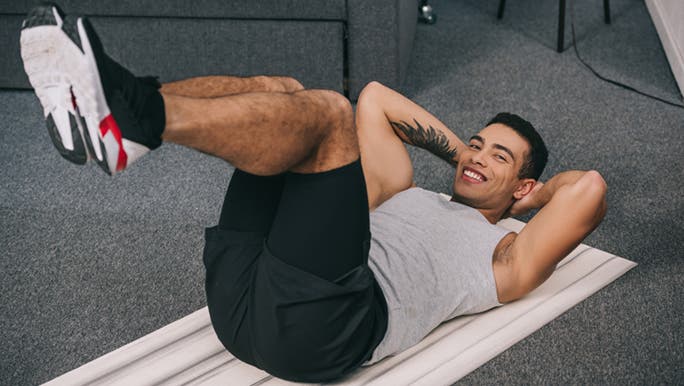 Doing crunches on his exercise mat in the lounge room, a young man smiles as he knows how to do exercise at home. 