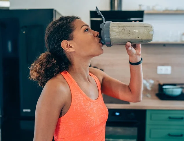 A woman in a bright orange racerback drinks the best pre workout powder from a protein shaker bottle.