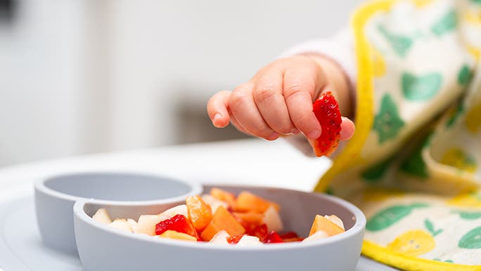 Up close, a baby is holding a strawberry in between their thumb and forefinger. There is a bowl of chopped fruit in front of them. 