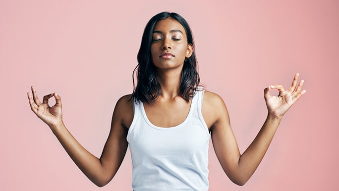 A woman meditates in front of a pale pink wall, her hands are up next to her and she is touching her thumb to her pointer fingers.