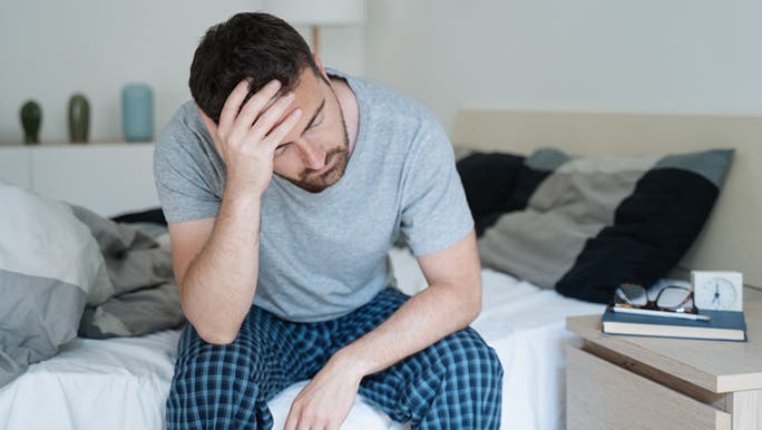  A man with dark hair wearing pyjamas is sitting on the edge of the bed with his head in his hand looking tired