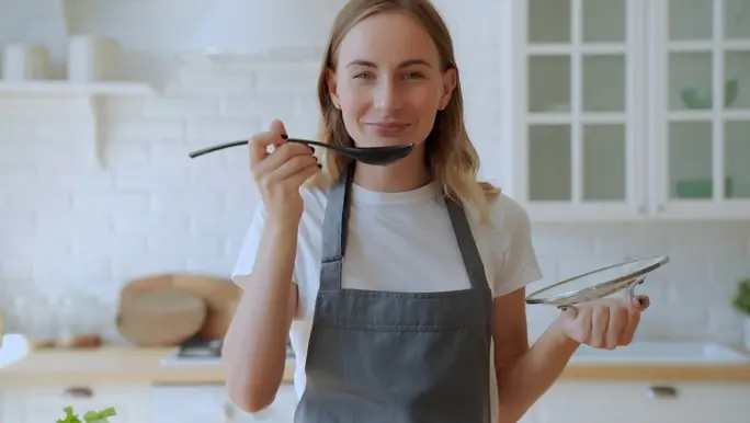 Woman cooking soup on stove at kitchen using pantry staples. Young woman tasting soup on spoon