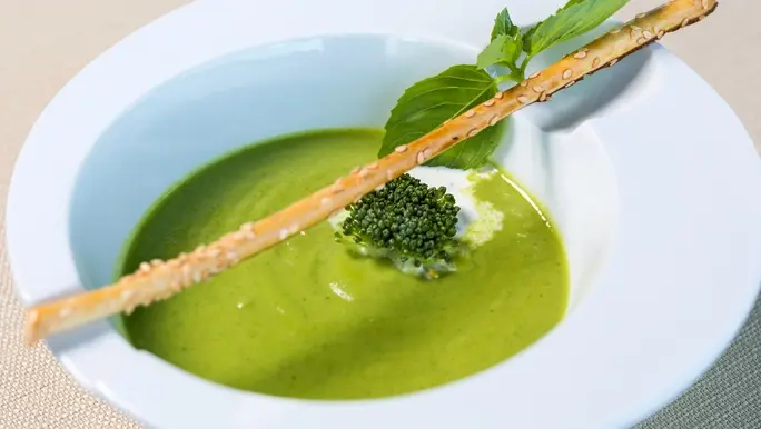 Spinach and broccoli soup in a white bowl with fresh basil and a bread stick
