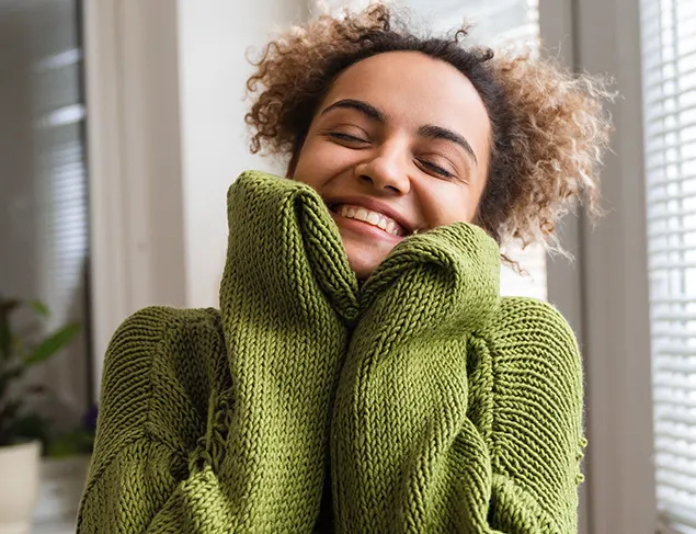 A woman in a soft green jumper smiles as she thinks about how to practise self-love. 