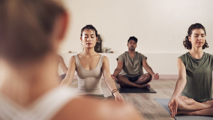 A group gym lesson is ending with meditation, three people are visible over the instructor's blurred shoulder.
