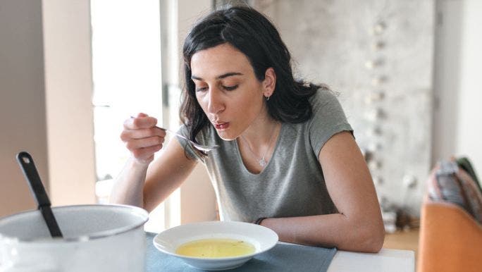A young woman with dark hair is sitting at a table eating a bowl of bone broth