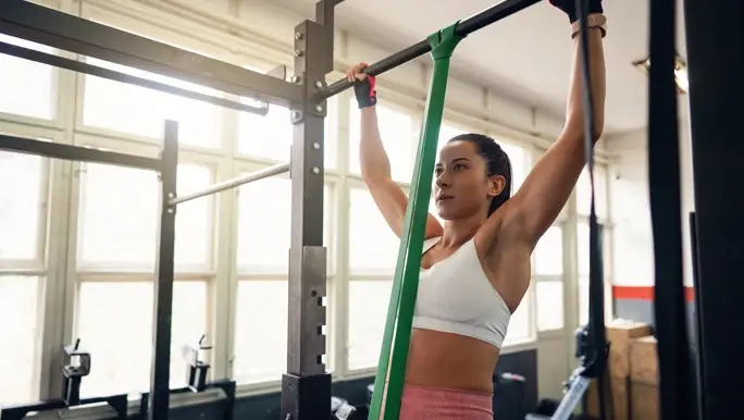 Woman in the gym doing a pull up assisted by a green resistance brand