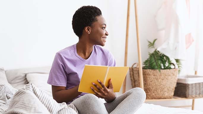 A woman sitting on a comfortable couch smiles while journaling which is part of her self-love practice for health.