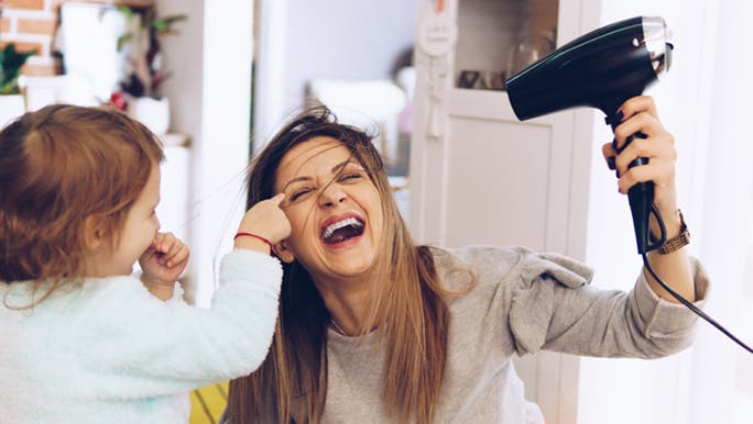Woman is blow drying her hair and laughing as her toddler points to her eye, she has beautiful healthy hair.