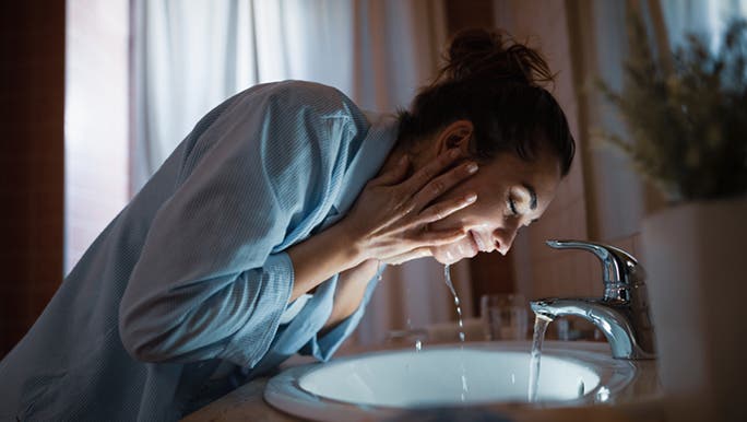 A lady is washing her face with both hands under the sink, as she’s splashing water onto her face, so she can wash off the cleanser that she uses to care for her oily skin at night. 