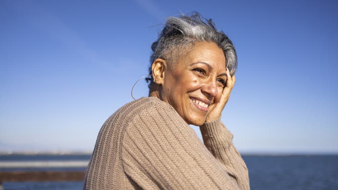 A woman smiles at the beach - the ocean is in the background and she has the look of someone who is aging and going grey gracefully.