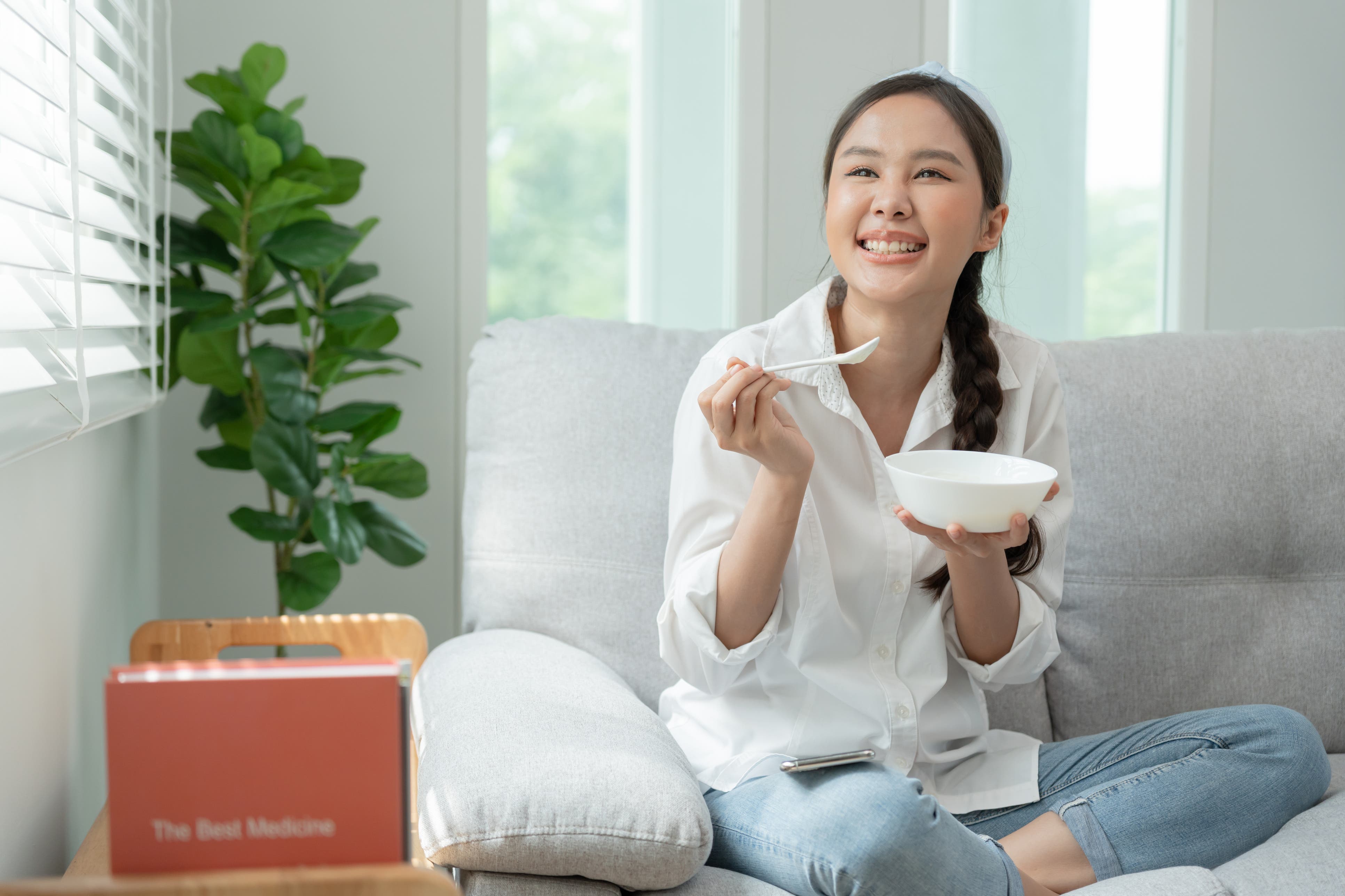 A young Asian woman is sitting cross-legged on a sofa eating from a white bowl