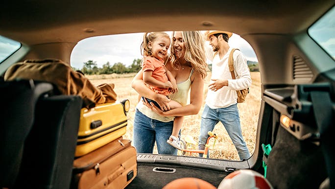From inside a car, the camera looks out an open boot past balls and suitcases to show a family in nature. 