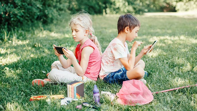 Two kids sit outside on the grass with their butterfly catching nets around them but they are both playing on their phones while you wonder how much screen time should kids have. 