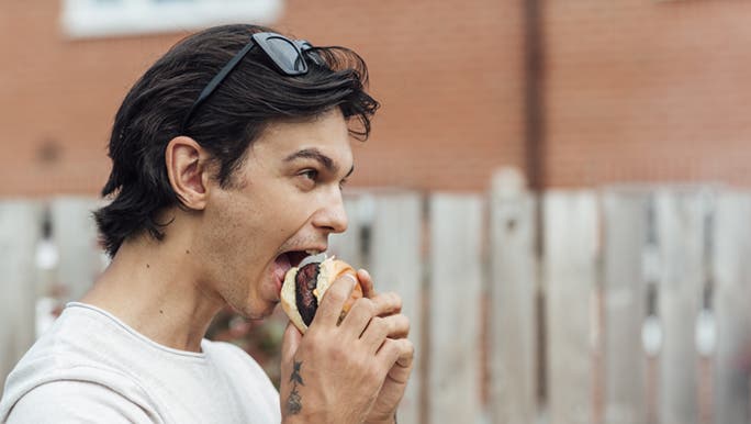 A man is standing outside biting into a plant-based burger. Perhaps, eating a plant based diet, is how you can eat meat sustainably.