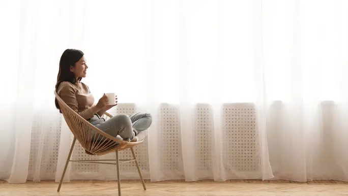 Relaxed woman sitting in a cane chair enjoying a cup of tea in front of a window with natural light