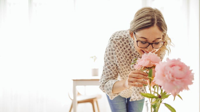Smiling blonde woman with glasses leaning in to smell fresh pink flowers in a vase