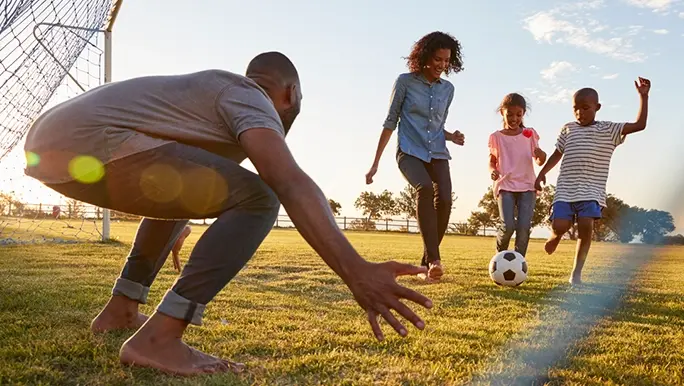 A family is playing soccer. The son is about to take a shot at goal and the dad is crouched down ready to try and save the ball.