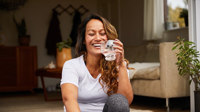 A woman smiles and drinks a glass of water while sitting on her front porch.