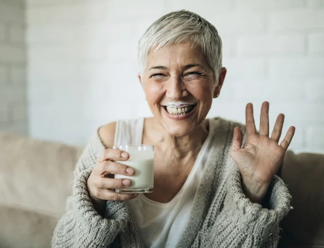 Happy woman with short grey hair drinking soy milk and laughing with a milk moustache