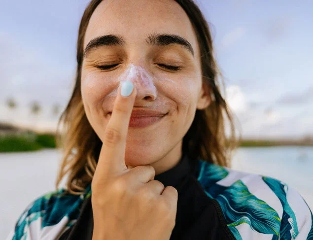 A woman in a sunshirt smiles as she puts sunscreen on her nose because she knows how sunscreen works. 