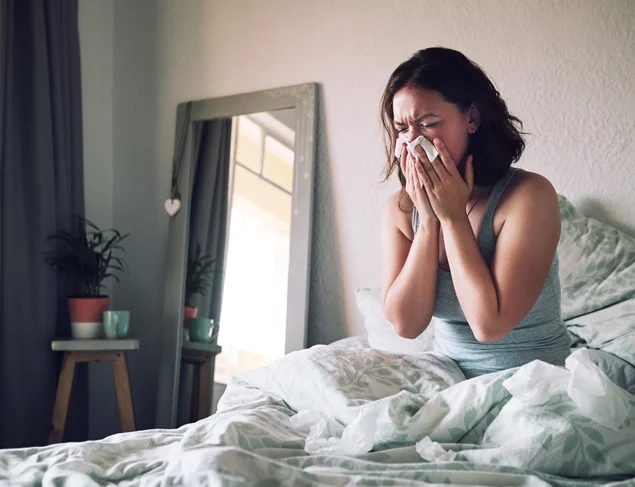 Woman suffering with the common cold and sneezing into a tissue while sitting on her bed at home