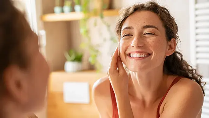 Woman smiles at herself in the mirror while thinking about practicing self love.
