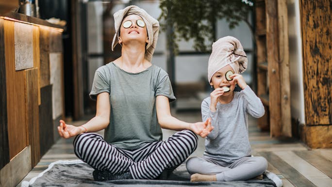 Sitting on grey timber floors, a mother and child meditate with towels on their heads and cucumber slices over their eyes as a self care strategy.