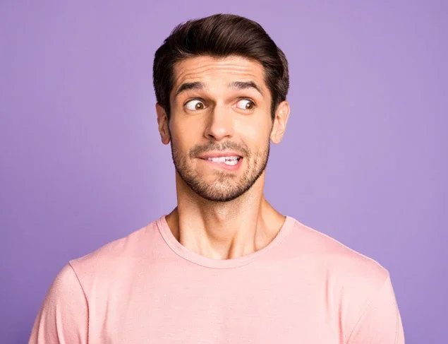 Man with a slightly embarrassed expression. He is wearing a pink t-shirt, standing in front of a purple background 